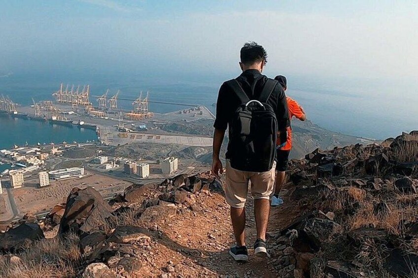 A person walking on a rocky path with a backpack, overlooking a coastal area with buildings and cranes. 5