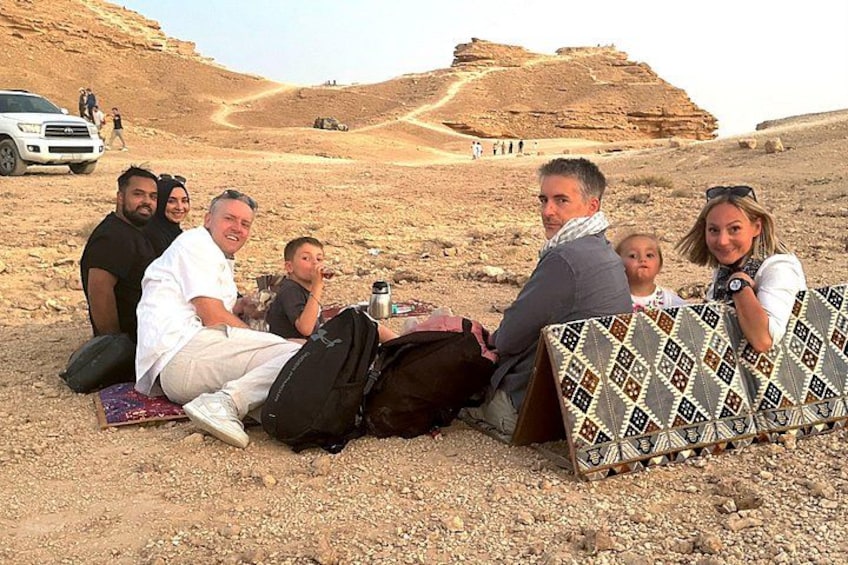 A group of people sitting on the ground in a desert landscape with rocky terrain and a large rock formation in the background. 4
