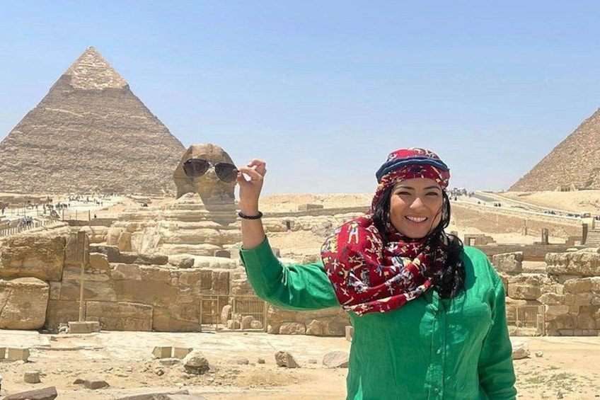 A person wearing a green shirt and a red patterned scarf, holding sunglasses in front of a pyramid. 4
