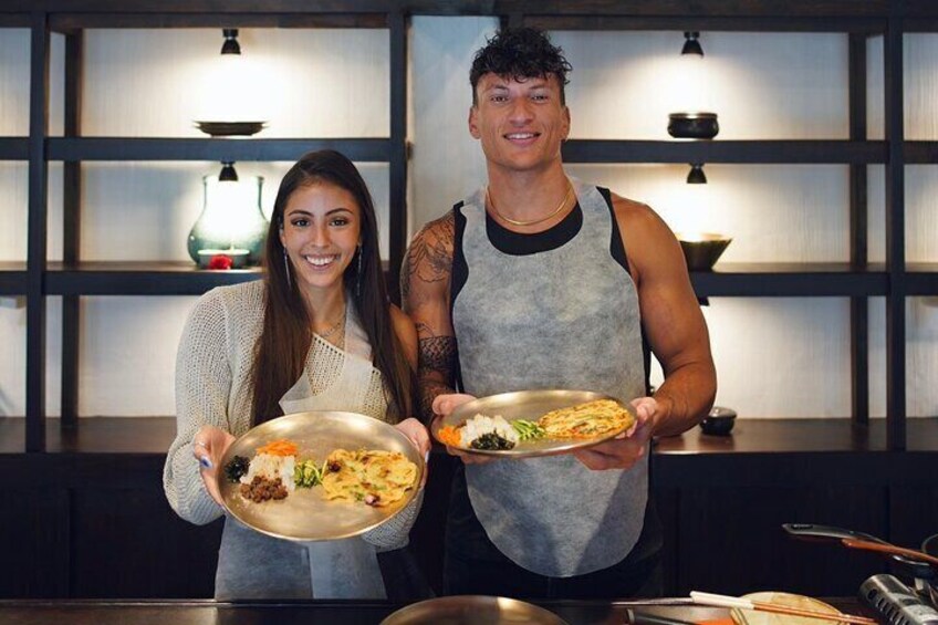 Two people holding plates of food in a kitchen with shelves and a lamp in the background. 3