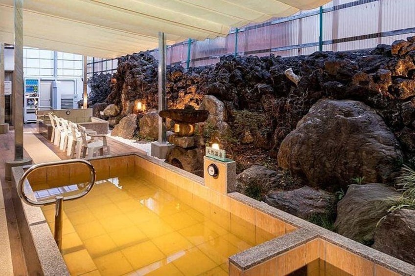 Un baño de aguas termales con un muro de roca de fondo, un banco de madera y un camino de piedra. 5