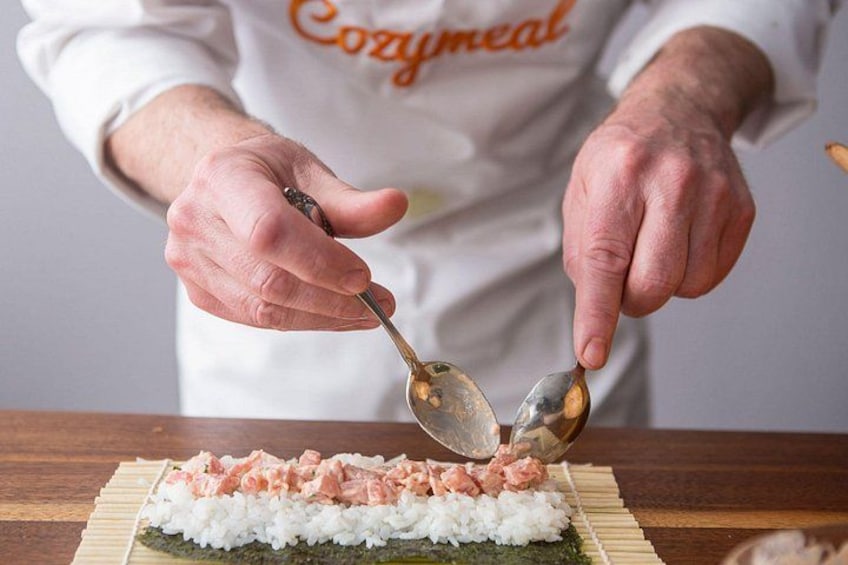 En person med ett vitt f örkläde med texten ”Cozymeal” på, som förbereder sushi genom att lägga ris och fisk på en bambumatta. 1