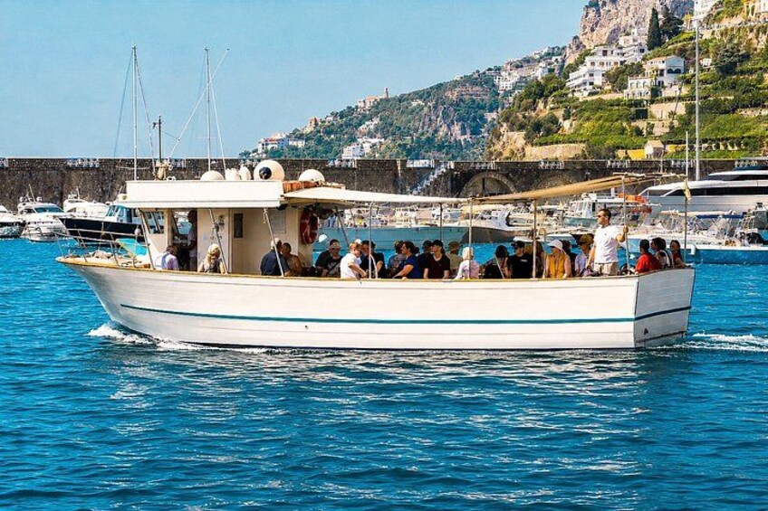 Een witte boot met passagiers, afgemeerd in een haven, met andere boten en een rotsachtige heuvelrug op de achtergrond. 3
