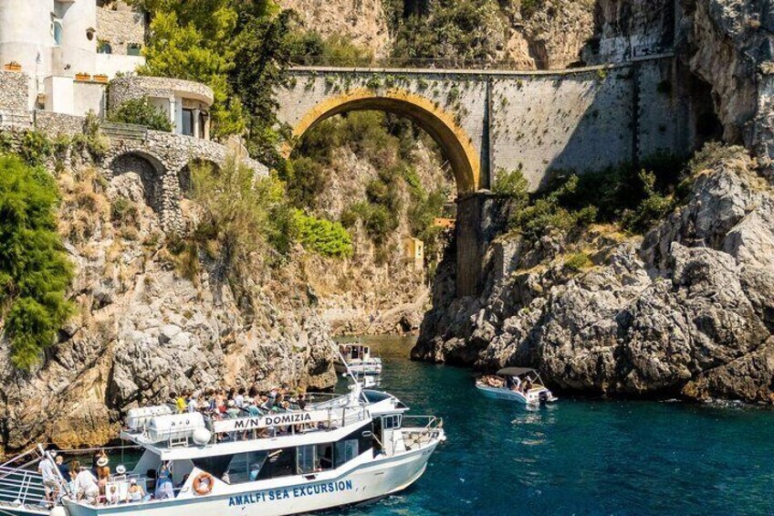 Un barco llamado "Amalfi Sea Excursion" atracado cerca de una costa rocosa con un puente de piedra al fondo. 5