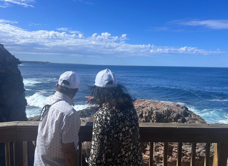 Two people overlooking a rocky coastline with waves crashing against the rocks under a clear blue sky. 2