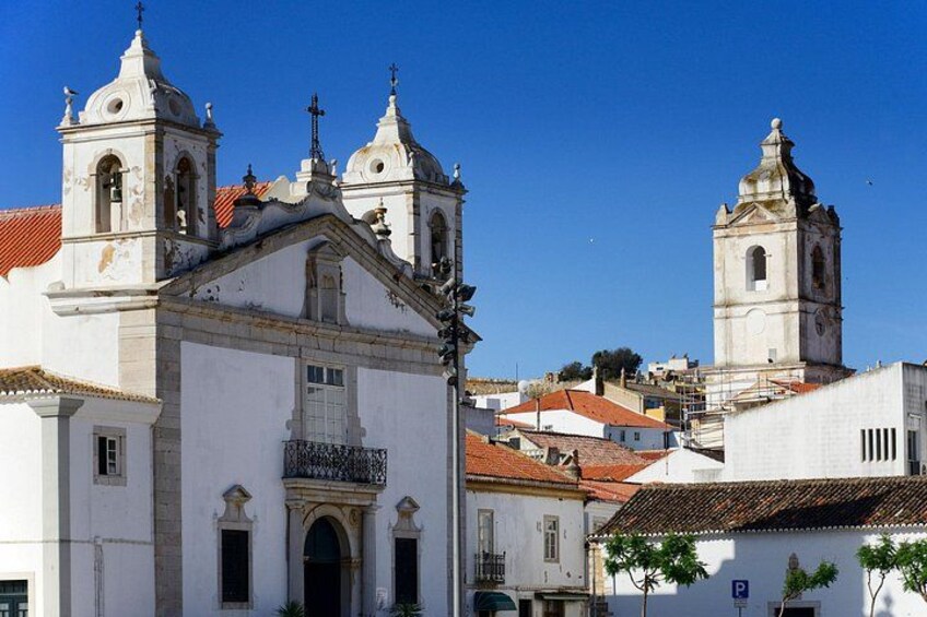 A white church with a bell tower and a smaller adjacent tower, surrounded by buildings with red-tiled roofs. 2