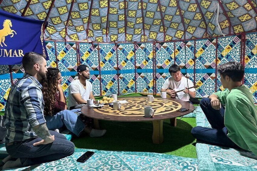 A group of people sitting around a circular table with a patterned tablecloth, under a tent with a colorful geometric design. 2