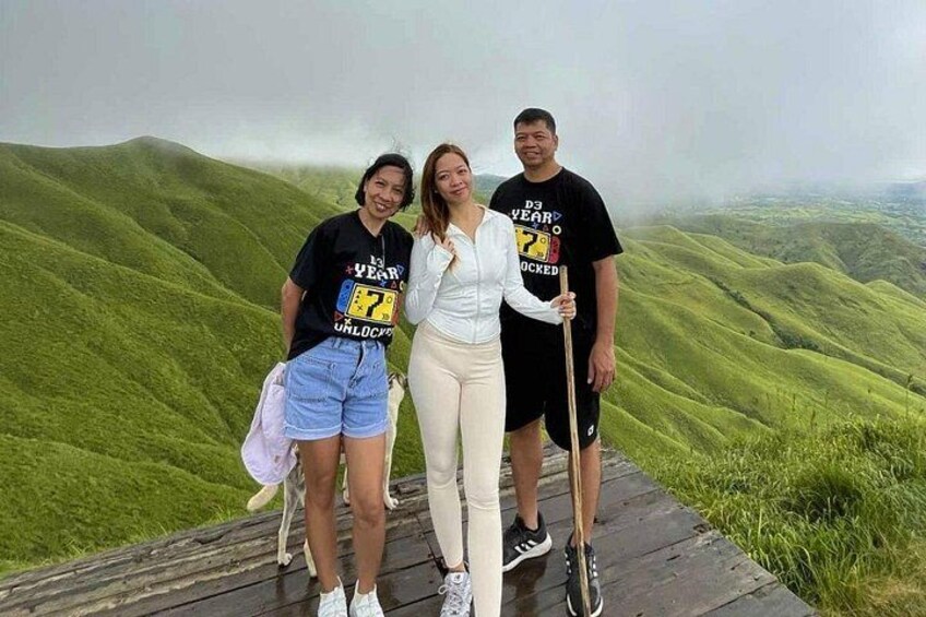 Three hikers pose on a wooden platform at Alicia Panoramic Park, surrounded by a stunning expanse of mist-covered rolling green hills. 1