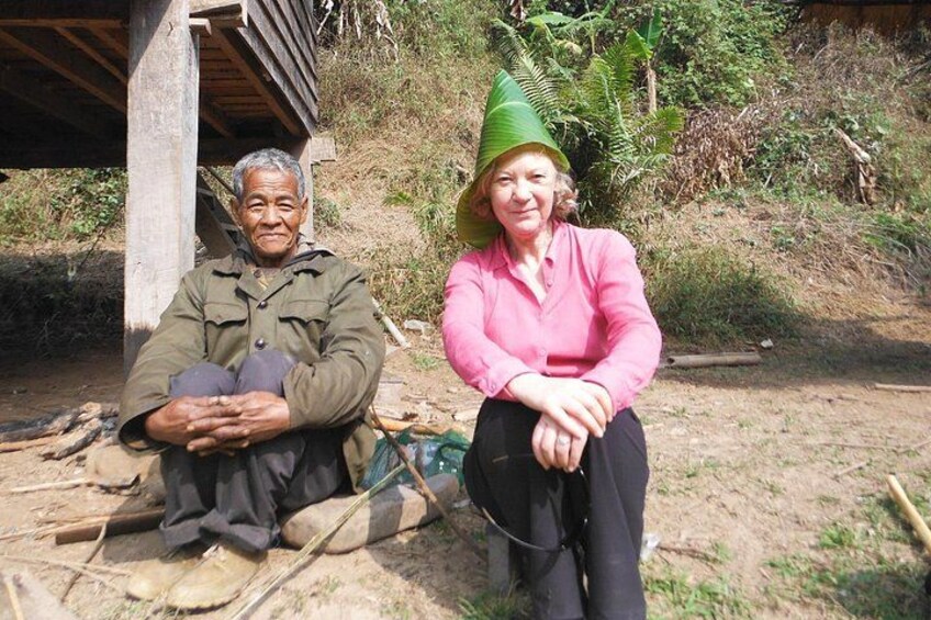 Two people sitting outdoors, one in a green jacket and the other in a pink shirt, with a wooden structure and greenery in the background. 4