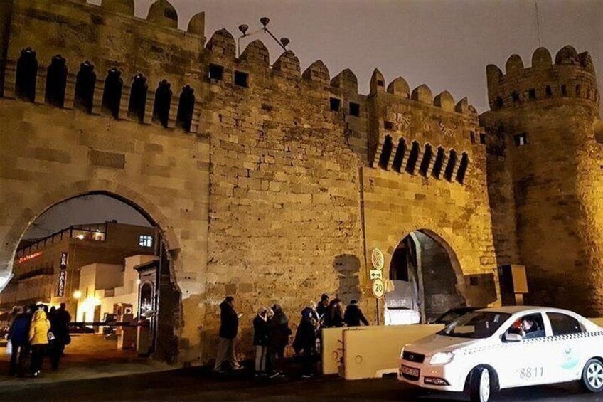 A medieval stone wall with arched gateways, a white car with a visible license plate, and a group of people standing near the wall. 4