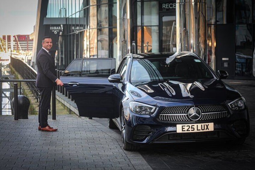 A man standing next to a black Mercedes-Benz E-Class car with a license plate E25 LUX. 2