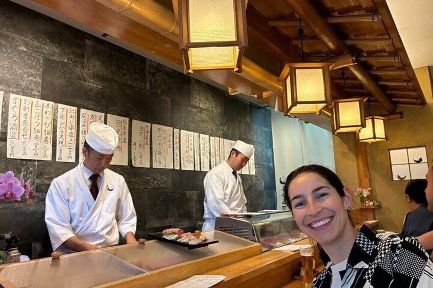 A sushi restaurant interior with chefs preparing food, wooden beams on the ceiling, and Japanese calligraphy on the wall. 1