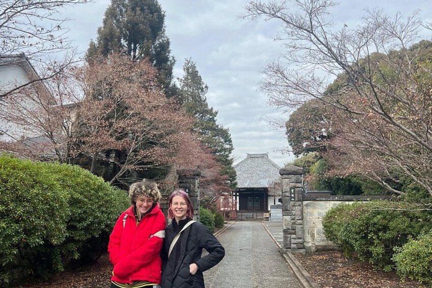 Two people standing in a garden with cherry blossoms, a stone pathway, and traditional architecture in the background. 4