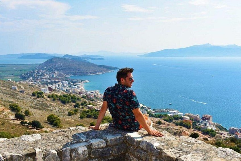 A person sitting on a stone wall overlooking a coastal town with a view of the sea and distant mountains. 3