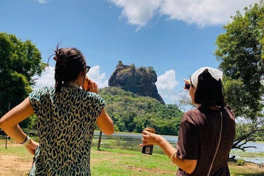 Two people standing by a lake, one holding a camera and the other holding a drink, with a large rock formation in the background. 1