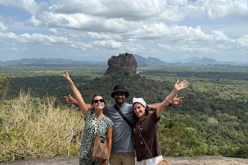 Three people standing on a hill with a view of a forested valley and a prominent rock formation in the distance. 2