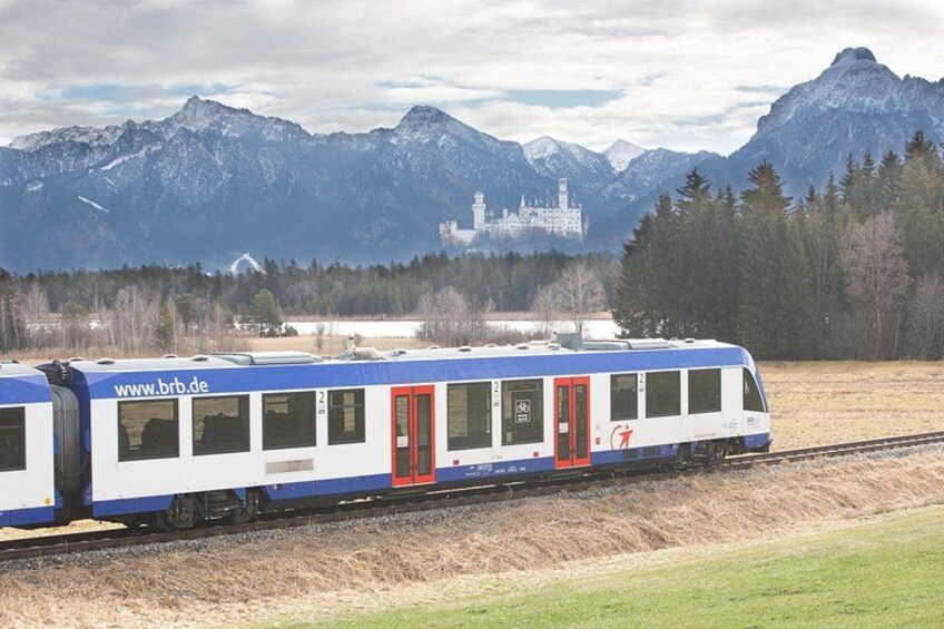 Un tren con puerta roja viajando sobre rieles, con un paisaje montañoso y un castillo al fondo. 5