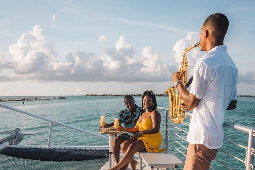A saxophonist playing on a boat with two people seated at a table. 1