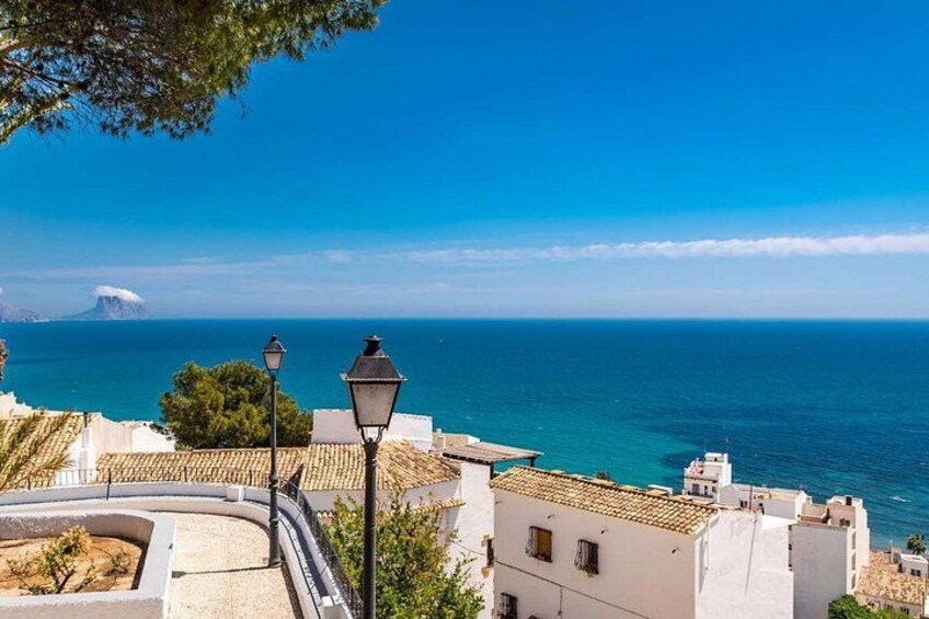A coastal town with white buildings, a curved road, and a view of the sea and a distant mountain. 3