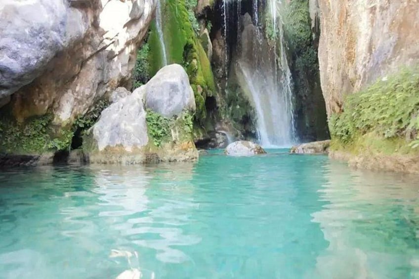 A clear turquoise pool with a waterfall, surrounded by large rocks and moss-covered cliffs. 2
