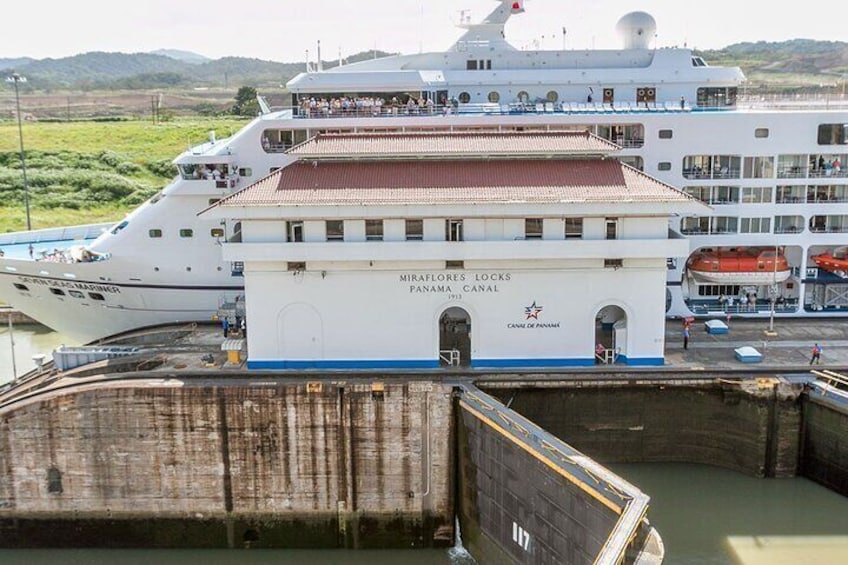 Een groot cruiseschip aangemeerd in een haven, met de naam "Miraflores Locks Panama Canal" zichtbaar op de zijkant. 1