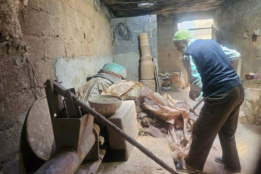 A person operating a manual food processor in a rustic kitchen with a concrete floor and walls, surrounded by various kitchen utensils and containers. 3