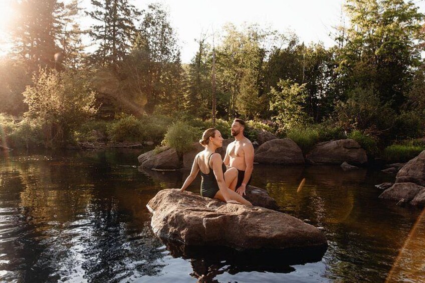 A couple sitting on a rock by a calm river surrounded by trees. 4