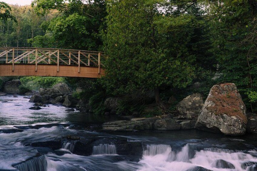 A wooden bridge over a flowing river, surrounded by dense greenery and large rocks. 2