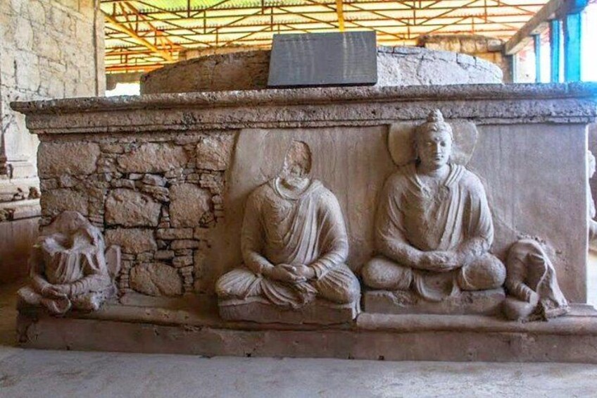 A stone sculpture of three seated figures in a meditative pose, with a stone wall and a metal structure in the background. 4