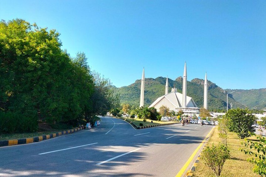 A road leading to a large mosque with four minarets, surrounded by greenery and hills. 3