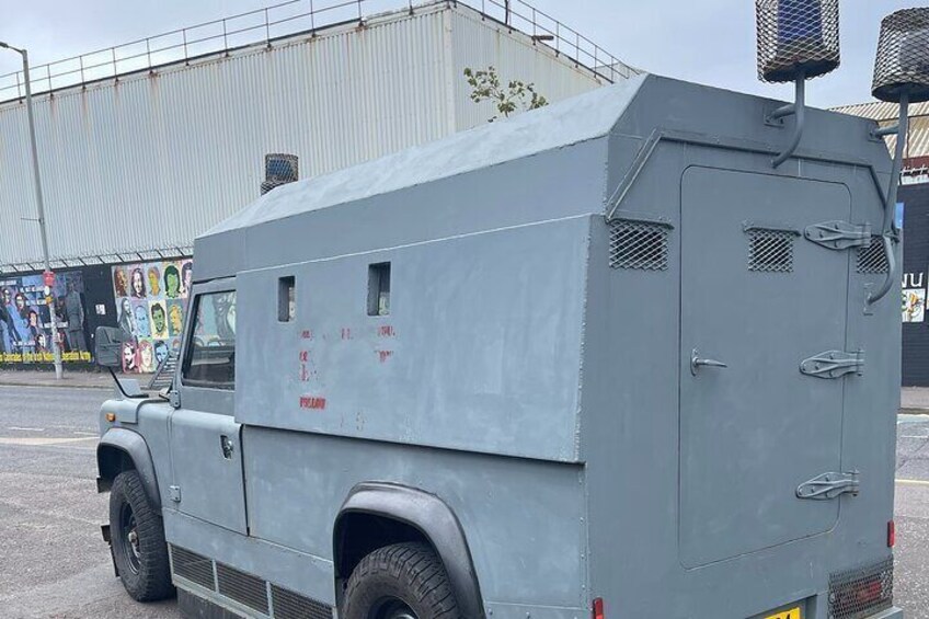 A gray military vehicle parked on a street with a mural in the background. 1