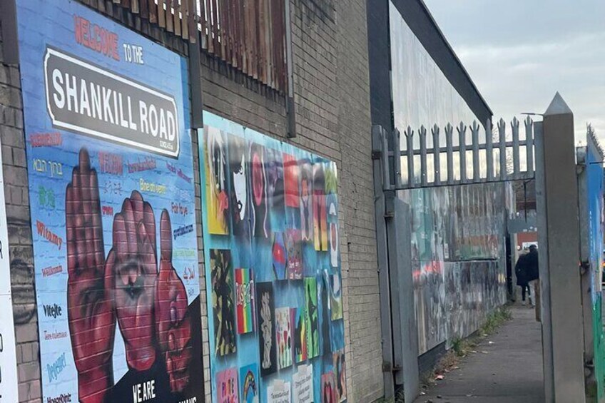 A street with a large sign reading "Welcome to the Shankill Road" and a gate with a fence. 3