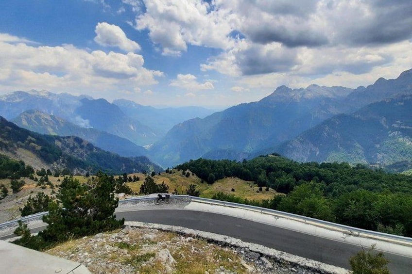 A winding mountain road with a guardrail, surrounded by lush green forests and distant mountain peaks under a partly cloudy sky. 2