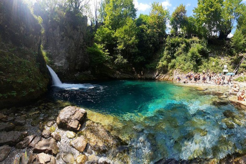 A clear blue pool with a waterfall, surrounded by greenery and rocks. 1