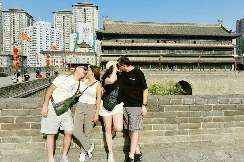 A group of people standing near a historical building with traditional architecture, a stone wall, and modern buildings in the background. 2