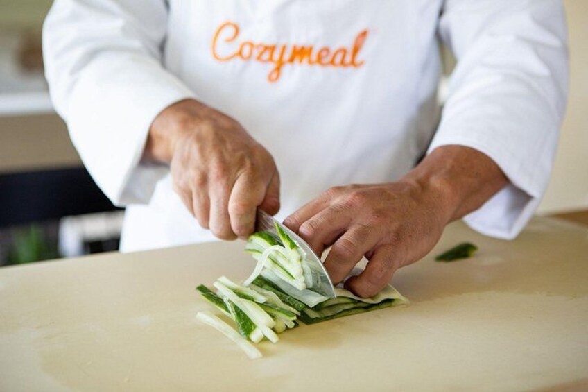 A person in a white chef's coat with 'Cozymeal' written on it, chopping green vegetables on a cutting board. 2
