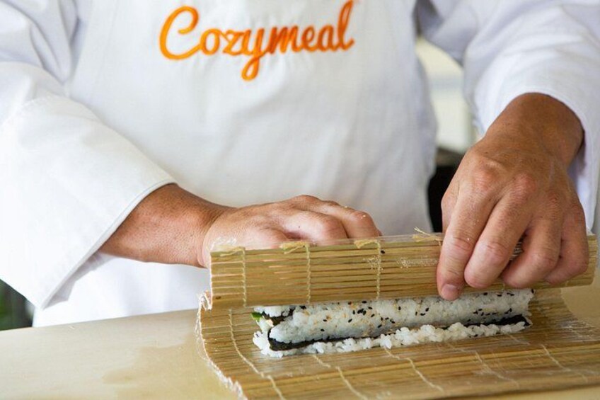 Eine Person in einem weißen Kochschürze mit dem Aufdruck 'Cozymmeal' rollt Sushi auf einer Bambusmatte. 1