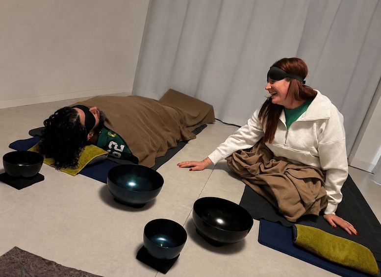 A person lying on a mat with a bowl, another person sitting beside them, and a window with blinds in the background. 4