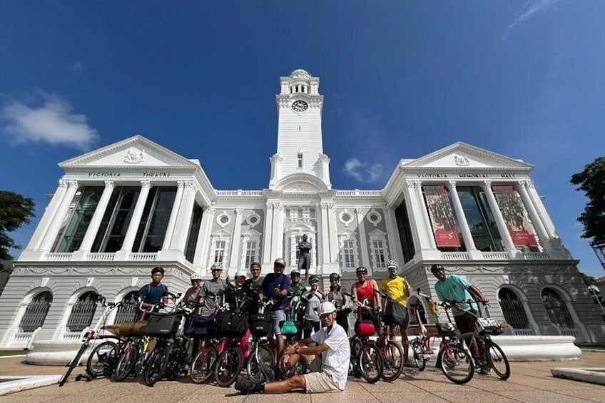 A group of people with bicycles in front of a white building with columns and a clock tower. 1