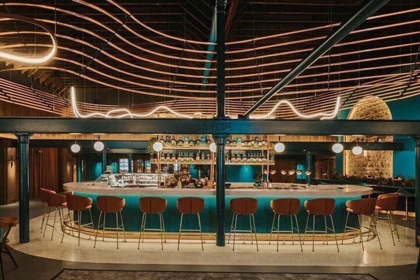 Een bar met een gebogen toonbank, houten stoelen en een plafond met zichtbare balken en verlichting. 2