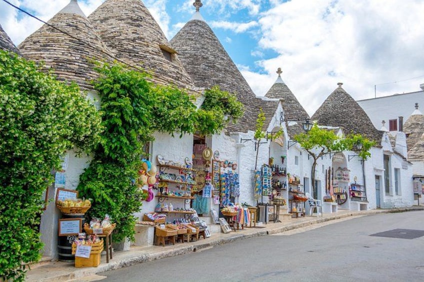 Una strada fiancheggiata da edifici bianchi con tetti a cono, negozi con articoli colorati e presenza di vegetazione. 4