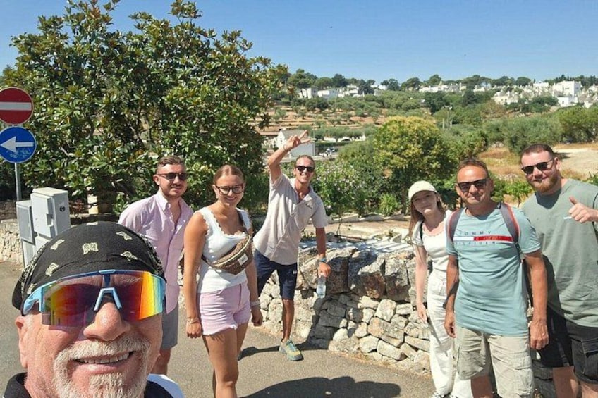 Un gruppo di persone in piedi su una strada, con un muro di pietra e vegetazione sullo sfondo. 3