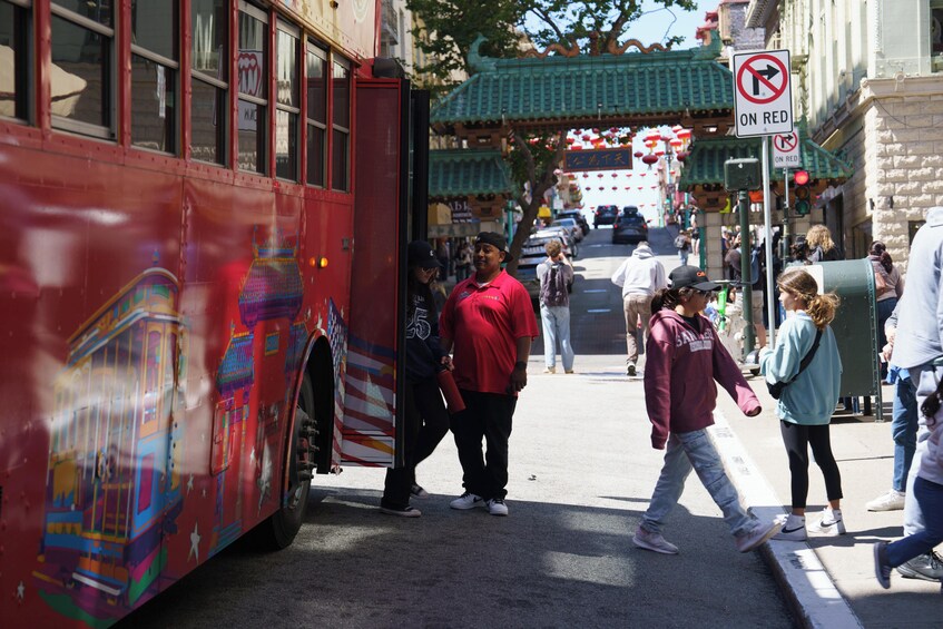 A red bus with colorful designs parked on a street. People are walking on the sidewalk, and there is a traditional Chinese gate in the background. 5
