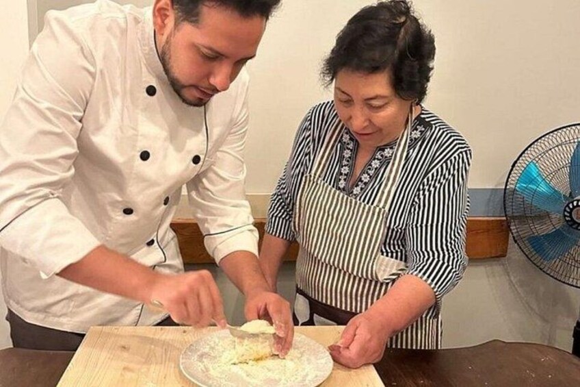 Two people preparing food, one in a chef's coat and the other in a striped shirt, with a fan in the background. 1