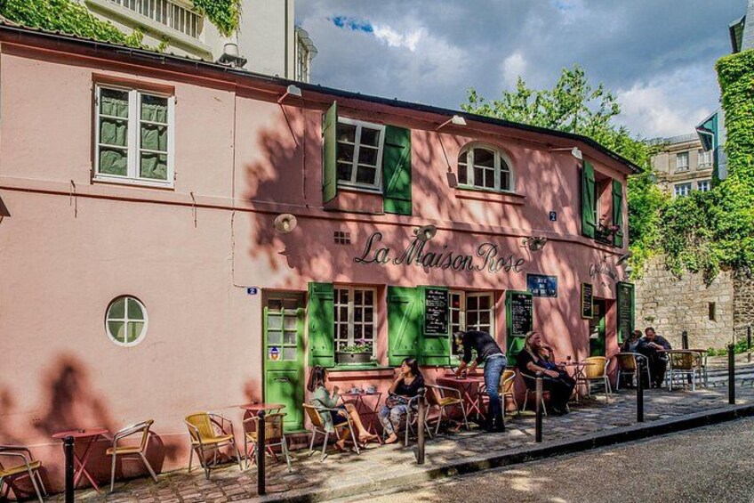 En rosa bygning med grønne skodder og navnet 'La Maison Rose' på facaden. Der er udendørs borde og stole, og folk sidder udenfor. 5
