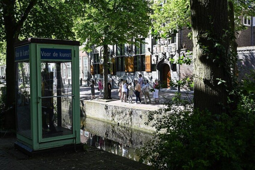 A green telephone booth with a sign reading 'Voor de kunst' in a public square with people walking around. 1