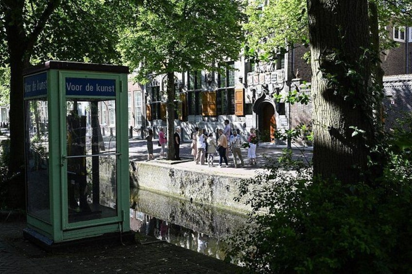 A green telephone booth with a sign reading 'Voor de kunst' in a park with trees and people. 1