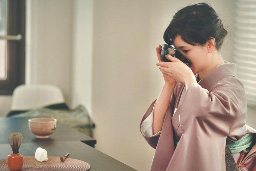 Una persona con una camicia rosa che tiene in mano una fotocamera, una ciotola e una frusta su un tavolo. 4