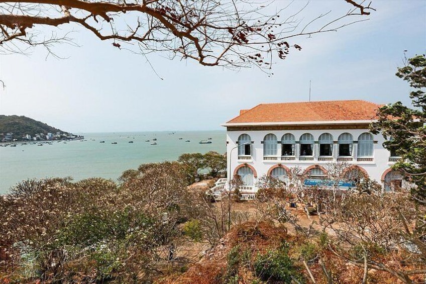 Un edificio costero con ventanas arqueadas, techo de tejas rojas y vista al mar con barcos. 4