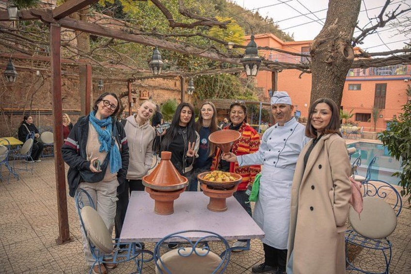 A group of people around a table with a clay pot and food, outdoors with a pergola and buildings in the background. 2